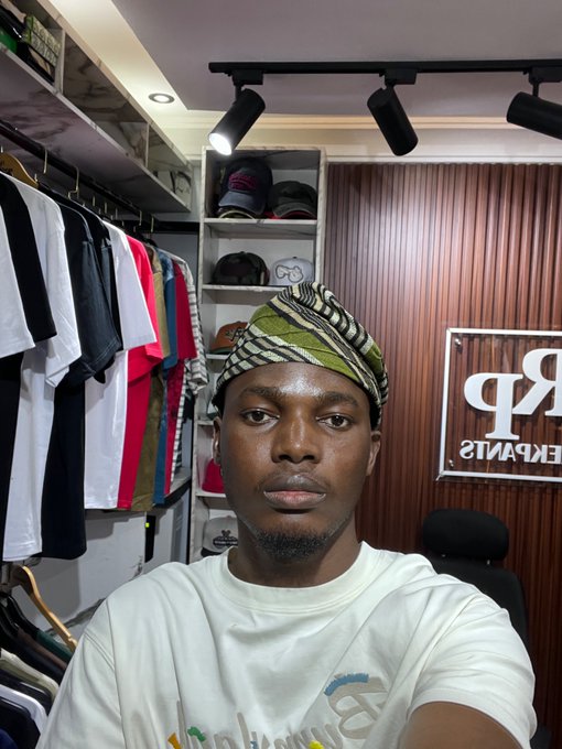A man wearing a patterned headwrap and white t-shirt takes a selfie in a clothing store with a 'РВ ЕКРАНИS' sign and shelves of caps in the background.