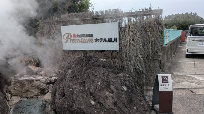 The image shows the entrance sign of "Premium Hotel 風月" (Shinzan Kanko Hotel) in Beppu, featuring a steaming hot spring and rock formations, with a smoking area sign nearby. This relates to the post about checking out of the hotel and heading to Beppu Station in the hot weather.