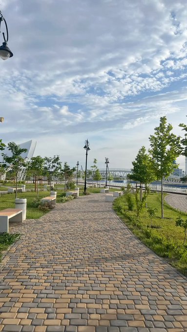 A paved walkway lined with young trees and benches leads through a park, with a bridge and stadium visible in the background under a partly cloudy sky.