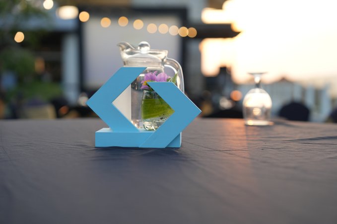 A light blue geometric napkin holder sits on a dark table alongside a glass pitcher of water and a glass, set at what appears to be an outdoor event.