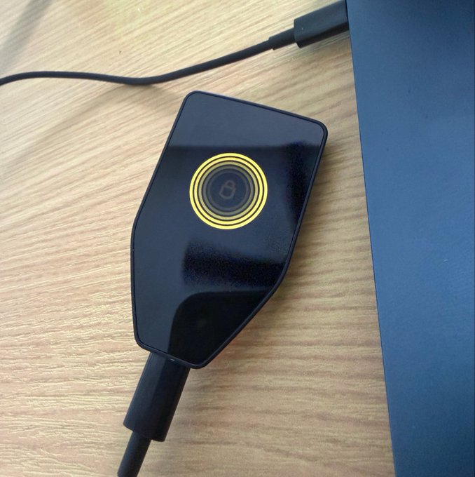A black wireless charging pad with a glowing yellow circular indicator and a logo, resting on a wooden surface next to a laptop and connected to a USB-C cable.
