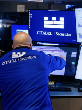 A photograph taken from behind showing a bald trader wearing a blue 'CITADEL | Securities' branded shirt, working at a trading desk with multiple monitors displaying the Citadel Securities logo on blue screens.