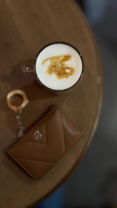The image features a wooden table with a cup of coffee, a keychain, and a wallet placed on top. The coffee appears to be latte-style, which has a caramel design on its surface. The keychain has a gold ring attached to it, and there is also a small purse present on the table. The post includes an image of a person's hand with their fingers spread out in front of them, accompanied by the text "GM Hug your coffee for a long day ahead Drifting Between Dreams".