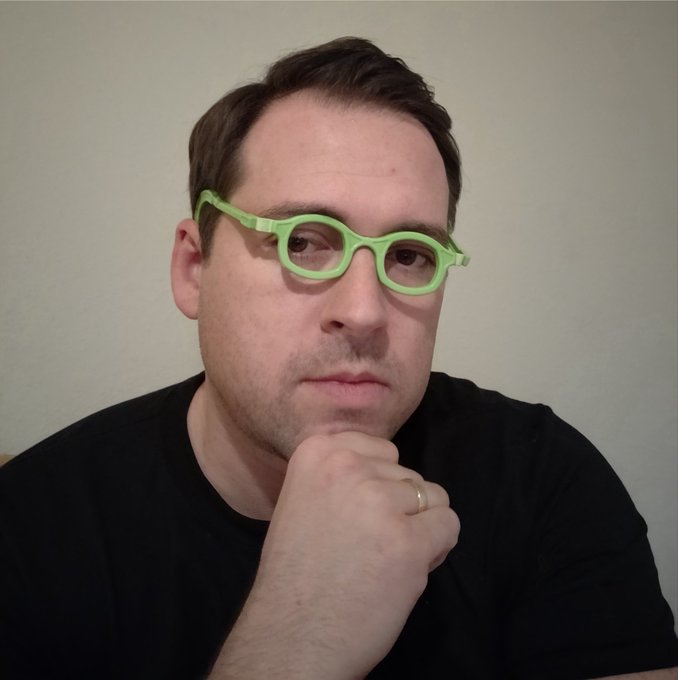A close-up portrait of a man wearing a black shirt and distinctive bright green glasses, resting his chin on his hand in a thoughtful pose.