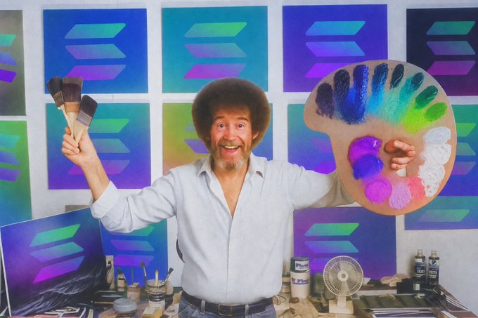 The artist Bob Ross standing in his studio holding paint brushes in one hand and a wooden palette loaded with vibrant colors in the other, smiling broadly with his iconic afro and beard, surrounded by his gradient geometric paintings.