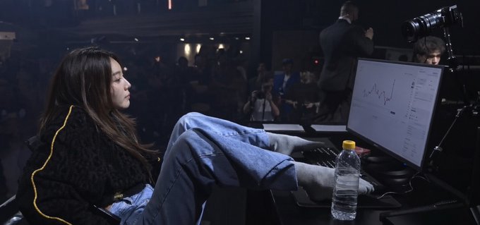 A young woman in a black jacket and jeans sits with her feet propped up in front of a computer monitor displaying financial charts, with photographers and a crowd visible behind her.