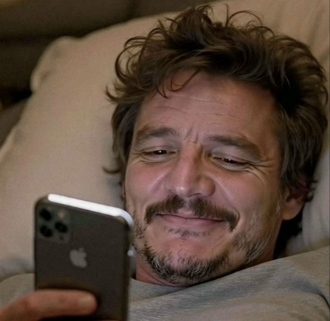 A man with curly brown hair, a mustache, and stubble lies in bed against a pillow, smiling warmly as he looks at a silver iPhone in his hand.