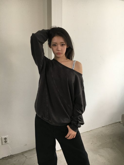 A portrait photo of a young woman wearing a dark gray off-shoulder sweatshirt with an Adidas strap visible, posing with one hand behind her head against a white wall.