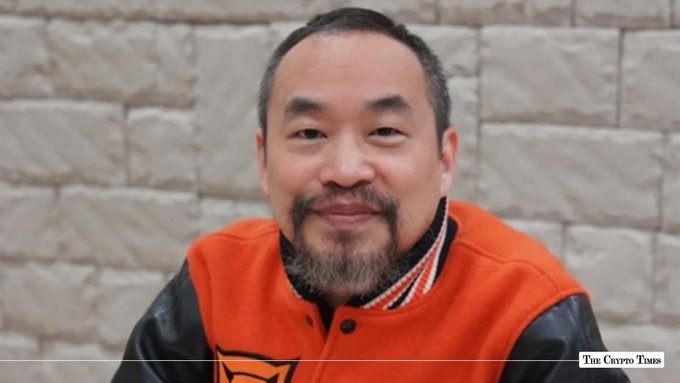 A man with a beard wears an orange and black varsity jacket, smiling in front of a stone wall, with 'The Crypto Times' watermark in the corner.