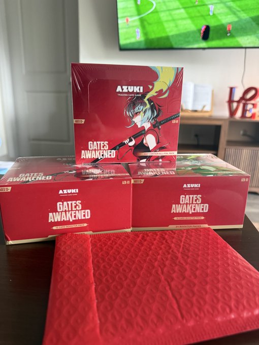 The image shows a collection of three red boxes, each containing a game called "Gates Awakened", placed on top of a red envelope or cardboard box. The boxes are neatly stacked together, with one box positioned above another and the third box below them. In the background, there is a TV screen displaying a soccer match, suggesting that these games might be related to sports or outdoor activities.
