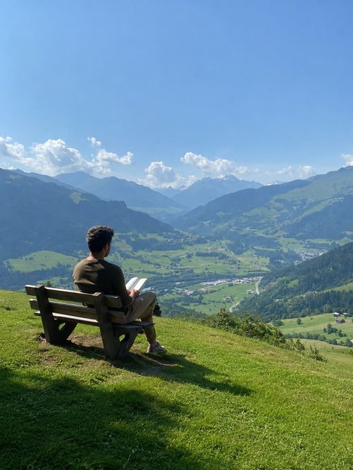 A person sitting on a wooden bench on a grassy hilltop, reading a book while overlooking a valley with mountains and a village in the distance under a blue sky.