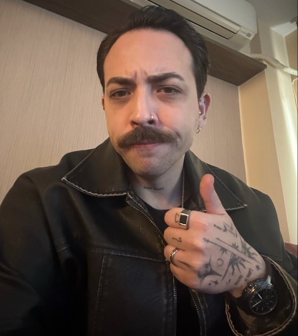 Selfie of a man with a mustache, nose ring, and tattooed hand giving a thumbs-up while wearing a black leather jacket and a large ring.