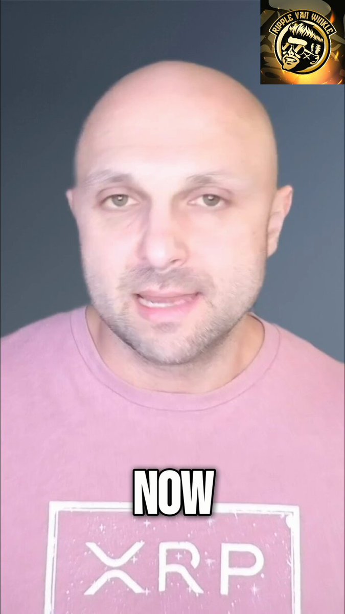 The image features a bald man wearing a pink shirt with white text that says "Now" and "XRP". The background of the photo is black, which contrasts with the man's pink shirt. There are no other objects or people visible in the image.