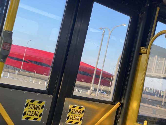 A view from inside a bus or train looks out through windows with yellow handrails and 'STAND CLEAR' warning signs, showing a red high-speed train passing by on an adjacent track under a clear blue sky.