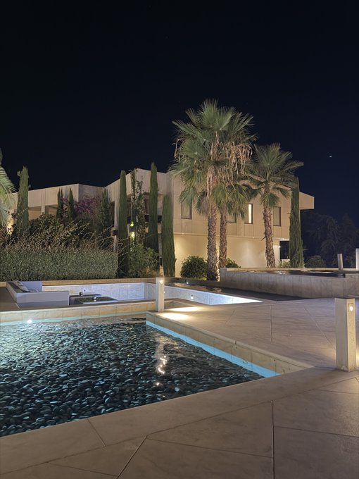 A nighttime photo of a luxurious backyard featuring a modern swimming pool, patio area with lounge furniture, tall palm trees, and a contemporary house illuminated in the background.