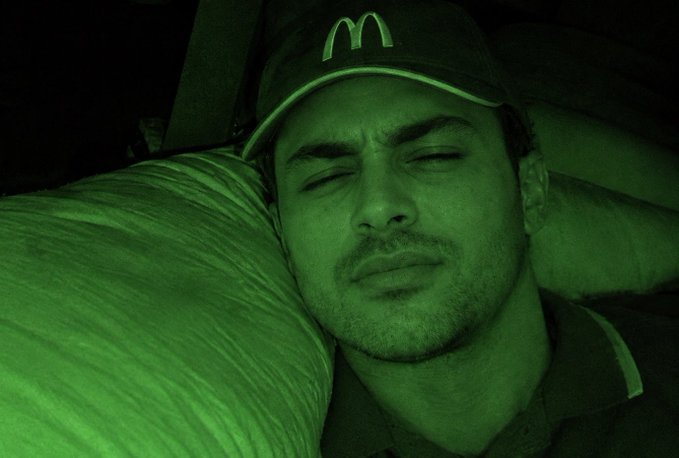 A man wearing a McDonald's cap sleeps against a striped pillow in what appears to be a vehicle, with the entire scene cast in an eerie green night-vision tint. The greenish glow emphasizes his groggy, just-woke-up expression, humorously matching his post about having a rough, nightmare-like experience before dozing off.