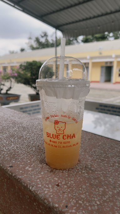 A clear plastic cup of 'BLUE CHA' iced tea with a dome lid sits on a speckled stone ledge, with text including 'Chanh - Tra Sua - Nuoc Ep - Sinh To' and a phone number.