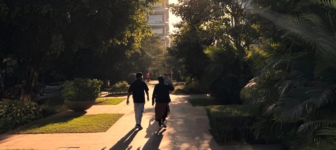 Two people walk down a sunlit, tree-lined paved pathway, with a tall building visible in the distance.