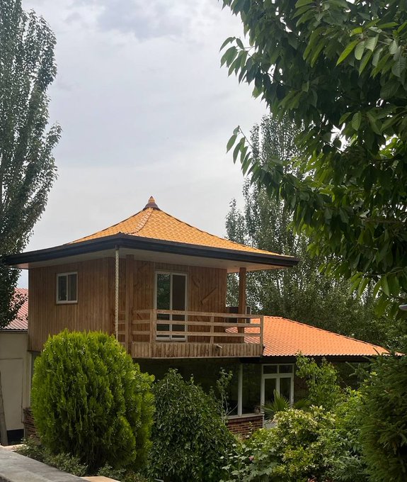 A two-story wooden house with an orange tiled roof, a wrap-around balcony, and surrounding lush greenery and trees under a hazy sky.
