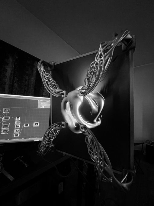 A high-contrast black and white photograph of a digital workstation showing a 3D model of an organic, alien-like creature with tentacle-like appendages displayed on a monitor, with node-based software visible on an adjacent screen.
