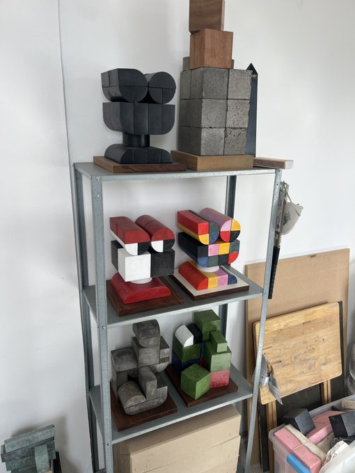 A metal shelving unit in a studio displaying various colorful geometric sculptures and wooden blocks, including stacked cubes in pink, white, green, and gray tones, with additional wooden panels leaning against the wall.