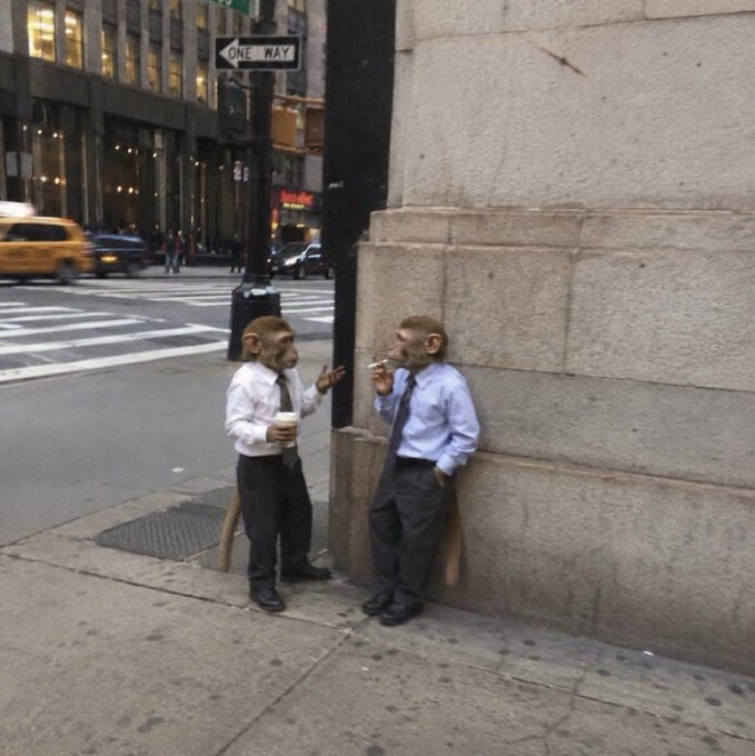 Two figures wearing monkey masks and business attire stand on a city sidewalk, taking a smoke break near a building corner with a 'ONE WAY' sign visible above.