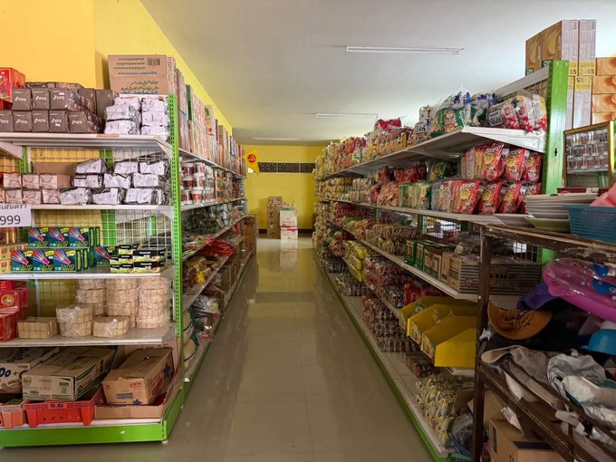 Interior view of a small grocery store with shelves stocked with various packaged goods, snacks, and household items on both sides of a narrow aisle.