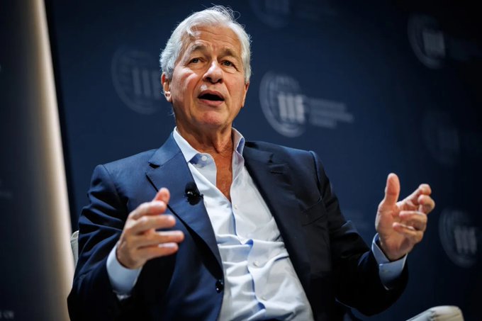 An older man with gray hair wearing a dark suit and open-collared light blue shirt speaks animatedly with both hands gesturing, set against a dark blue background with repeating logos.