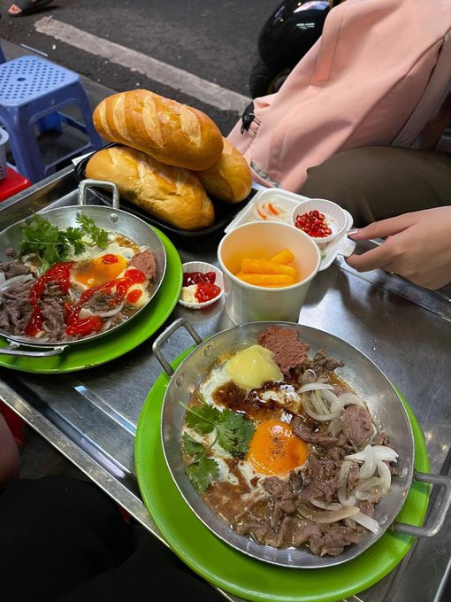 An overhead shot of a Vietnamese street food meal shows two metal skillets of sizzling beef and eggs on green plates, accompanied by baguette sandwiches, dipping sauces, and mango slices on a metal table.