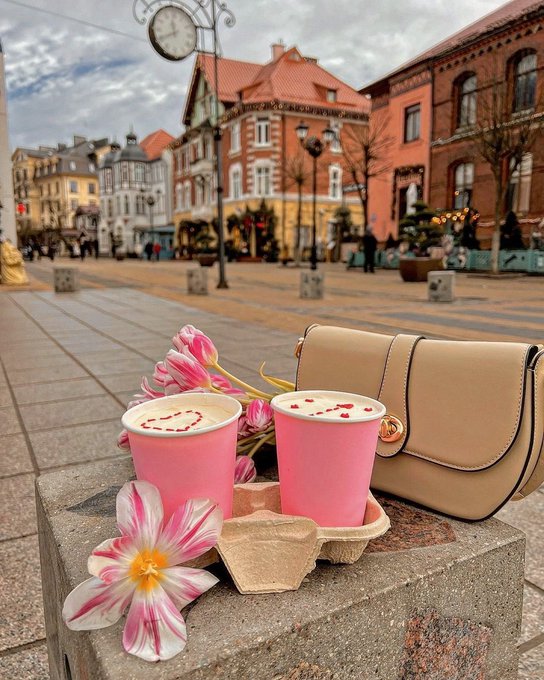 Two pink beverages with heart latte art, pink tulips, and a beige handbag arranged on a stone block in a European city square with a clock tower and colorful buildings.