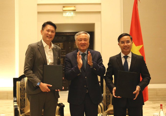 Three men in suits pose during what appears to be a formal signing ceremony, with two holding black document folders and the Vietnamese flag visible in the background.