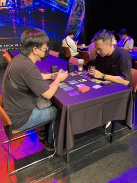 Two people seated at a table at a gaming or TCG event, appearing to play a card game together.