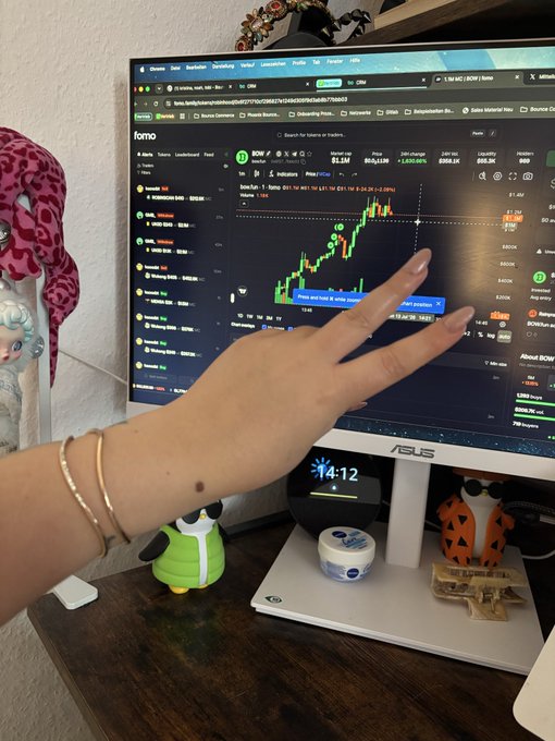 A person's hand making a peace sign in front of an ASUS computer monitor displaying a crypto trading chart on dexscreener.com for the token 'fomo' (SOUL), with small figurines on the desk below.