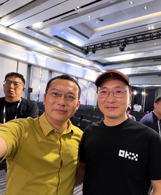 Two men posing for a selfie at what appears to be a conference or event venue, one wearing a yellow shirt and the other wearing a black t-shirt with 'OKX' branding and a cap.
