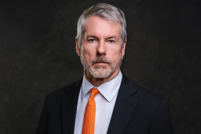 A professional portrait of a gray-haired, bearded man in a dark suit and orange tie against a dark background.
