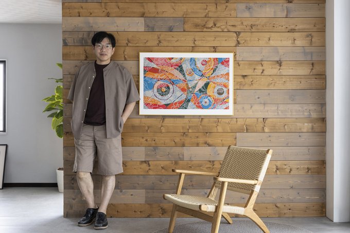 A man wearing glasses, a gray short-sleeved shirt, and shorts stands next to a wooden chair in front of a wood-paneled wall displaying a colorful framed artwork.