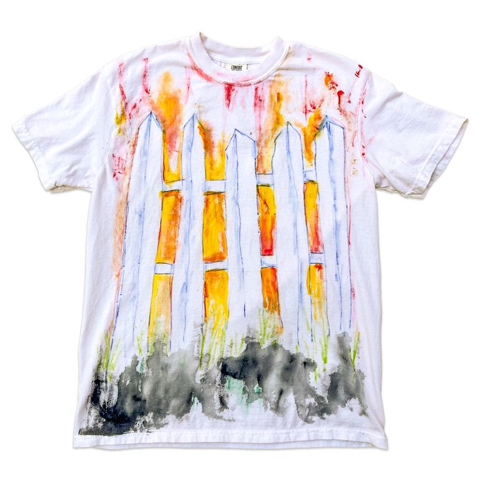 A white short-sleeve t-shirt features a hand-painted, abstract design of four vertical towers rendered in orange, yellow, red, and blue with flames and smoke at the base, resembling the World Trade Center.