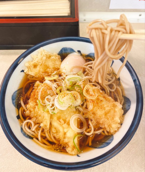 The image features a bowl of noodles with meat, vegetables, and egg yolks sitting on top of a table. The noodles are cooked and appear to be served in a blue and white bowl. A fork can also be seen next to the bowl, ready for use when eating the dish.