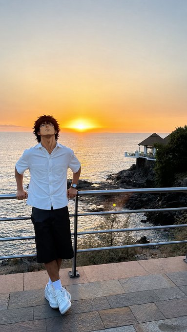 An anime-style filter is applied to a man wearing a white button-up shirt, black shorts, and white sneakers, leaning against a railing overlooking the ocean at sunset.