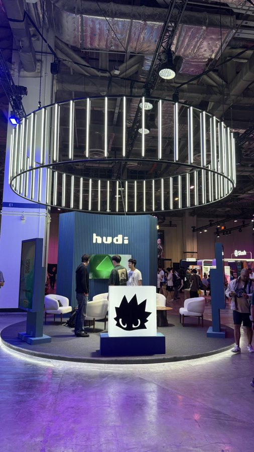 An exhibition booth for "hudi" featuring a large circular hanging light installation and a black-and-white mascot face logo on display.