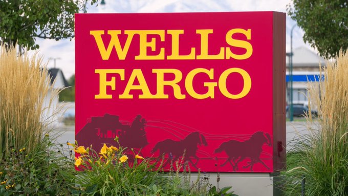 A red Wells Fargo sign with yellow lettering and a stagecoach silhouette sits amid landscaping. The image illustrates the post about the bank's $2.2 trillion asset institution planning to introduce tokenized deposits for corporate clients.