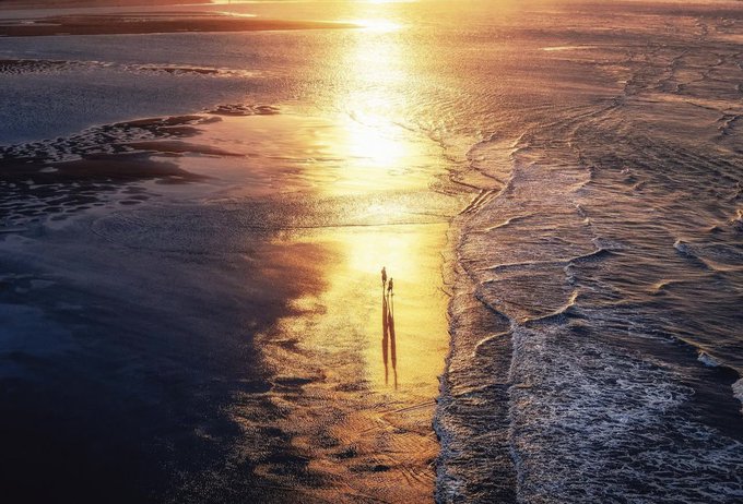 An aerial sunset view of a beach where two silhouetted figures walk along the wet sand, their long shadows cast by the golden sunlight reflecting across the water's edge.