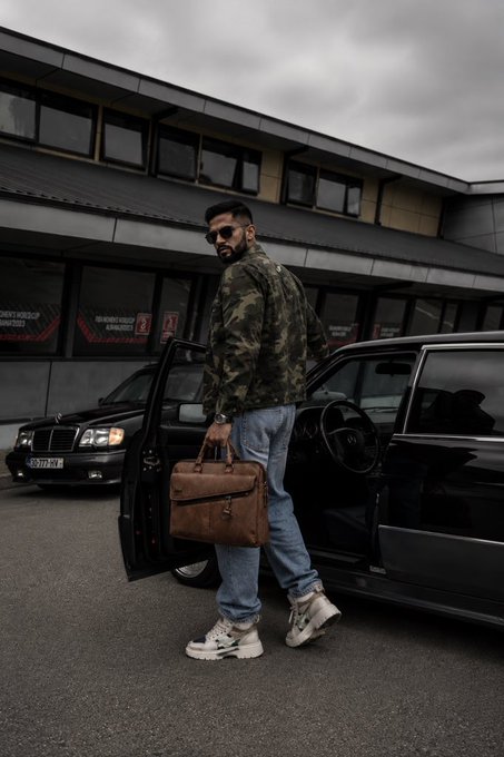 A man in a camo jacket, jeans, and chunky white sneakers holding a brown leather bag while stepping out of a black luxury car.
