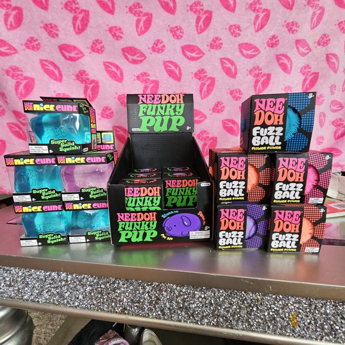 A display of toy products on a counter against a pink lips-patterned background, including boxes labeled "Nice Cube Sugidoll Squish", "NeeDoh Funky Pup", and "NeeDoh Fuzz Ball Fuzzee Power".