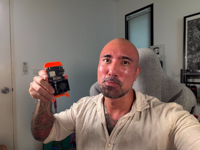 The image shows a bald man holding up an orange cell phone that has a camera attached, possibly for taking pictures of himself or his surroundings. The man appears to be wearing a white shirt and is sitting down while holding the phone. There are no other people in the scene, and the focus seems to be on the man with the cell phone.