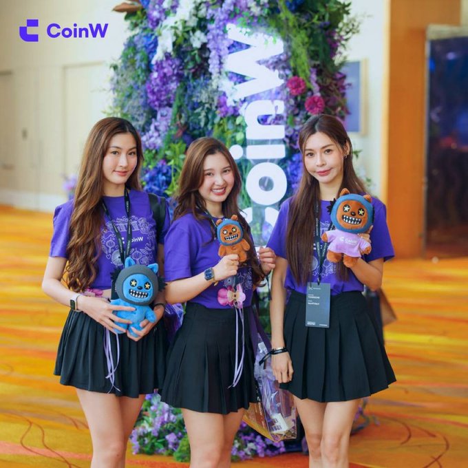 Three young women in matching purple CoinW t-shirts and black pleated skirts pose together holding plush toys in front of a floral display bearing the CoinW logo.