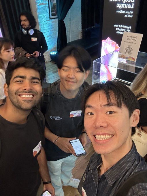 Three young men smile for a selfie at what appears to be a lounge or event space, with name tags visible and a partially reversed sign reading 'Lounge' and 'Reserved' in the background.
