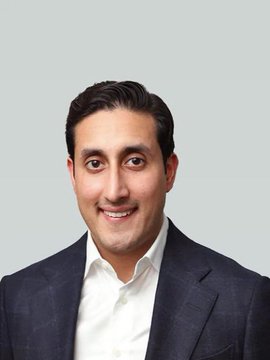The image features a man wearing a suit, smiling at the camera with his eyes closed. He appears to be posing for the photo, possibly as part of an advertisement or promotional material. The background is plain white, which helps draw attention to the subject in the foreground and emphasizes the main focus on the man's face and attire.