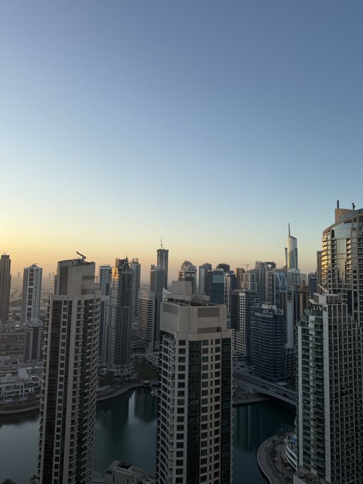 A scenic aerial photograph of a city skyline at sunset, featuring numerous modern skyscrapers and a calm body of water reflecting the buildings under a clear sky transitioning from blue to orange.