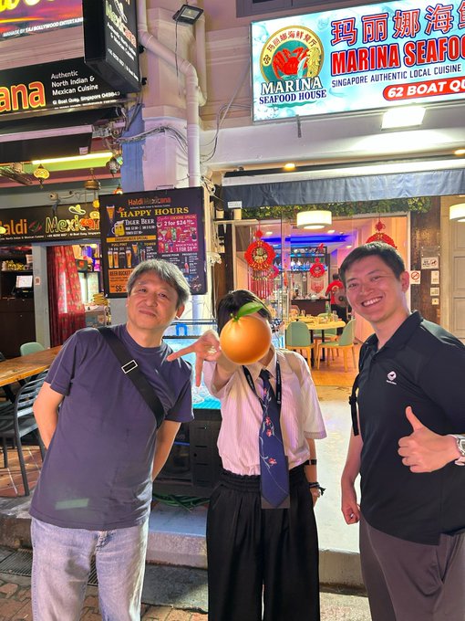 Two men pose for a photo with a third person who has their face covered by an orange fruit emoji, standing in front of the 'Marina Seafood House' restaurant, which displays signs for 'Authentic North Indian & Mexican Cuisine' and 'Happy Hours'.